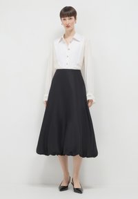 Frau in einer weißen Bluse mit langen Ärmeln und Knopfverschluss, kombiniert mit einem schwarzen, hochtaillierten Midi-Rock mit Ballon-Saum und schwarzen spitzen Slingback-Schuhen.