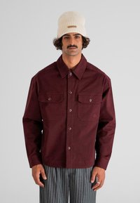 Hombre con un gorro de punto color crema, chaqueta burdeos de botones con bolsillos en el pecho y pantalones grises de rayas, de pie contra un fondo plano.