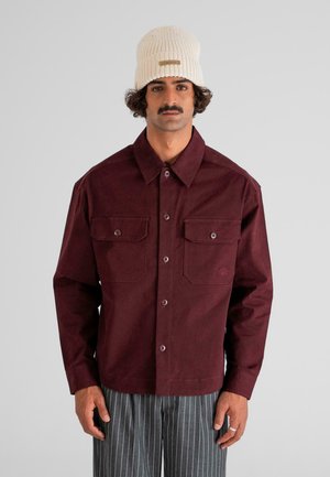 Man wearing a cream knit beanie, burgundy button-up jacket with chest pockets, and gray pinstripe trousers, standing against a plain background.