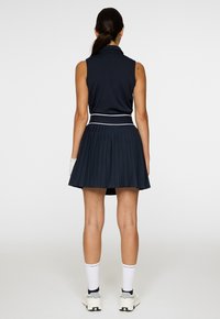 Nainen seisoo selkä kameraan päin, päällään ilman hihoja oleva tummansininen pusero, laskostettu hame, valkoiset sukat, lenkkarit ja vasemmassa kädessä yksi valkoinen käsine.