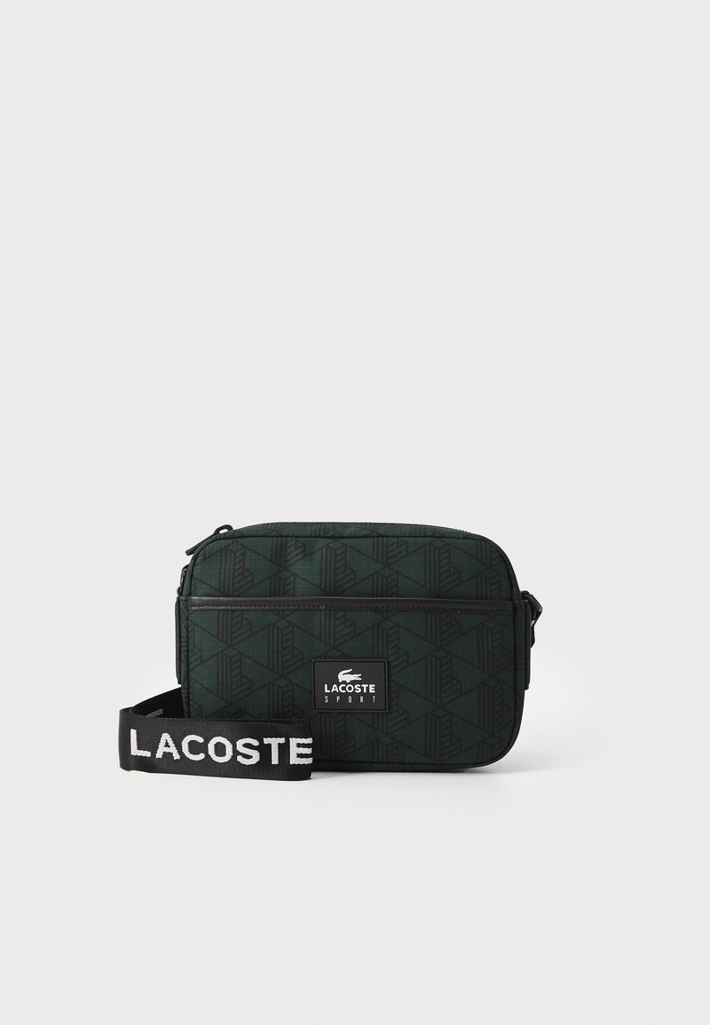 Borsa a spalla verde con un motivo a fantasia, di forma rettangolare, realizzata in tessuto testurizzato, con una cerniera nera e una tracolla staccabile con lettering bianco.