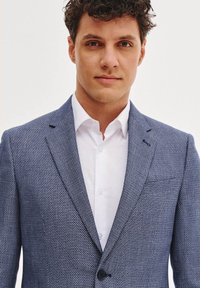 Niebieska żakiet o teksturowanej strukturze, dopasowany fason, z jedynym zapięciem na guzik i białą koszulą pod spodem.