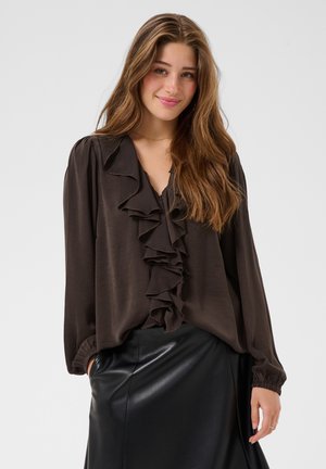 Jeune femme aux longs cheveux bruns portant un chemisier brun foncé à volants et une jupe en cuir noire, souriant doucement sur un fond uni.