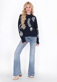 Eine Frau mit langen blonden Haaren trägt einen marineblauen Pullover mit weißen Blumenmustern, hellblaue Schlagjeans und beigefarbene Boots. Sie steht selbstbewusst.