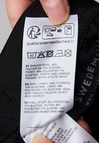 Svart tygvårdsetikett som visar tvättanvisningar och materialkomposition: 90% polyester, 10% elastan, översättningar på flera språk.