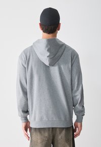 Grå hettegenser laget av myk tekstil, med stor hette og ribbestrikkede mansjetter, sammen med mørkfargede bukser, sett bakfra.