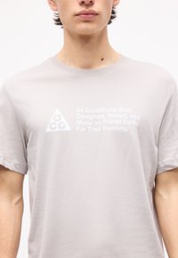 Νεαρό άτομο που φοράει ανοιχτό γκρι μπλουζάκι με λευκό κείμενο "Εξοπλισμός για όλες τις συνθήκες, Σχεδιασμένος, Δοκιμασμένος και Κατασκευασμένος στον πλανήτη Γη, για ορεινό τρέξιμο."