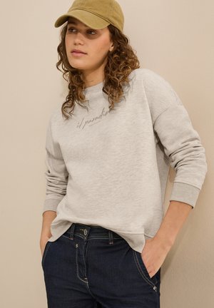 Hellgrau Sweatshirt mit gerippten Bündchen, bedruckt mit dem Text "il paradiso positivity". Kombiniert mit dunklen Jeans und einer khakifarbenen Mütze.