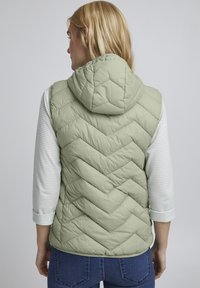 Hellgrüße, gestepptes Weste mit Kapuze, die ein Chevron-Muster aufweist, und einem weißen, strukturierten Langarmtop darunter.