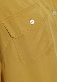 Chemise jaune avec une poche sur le côté gauche de la poitrine, fermeture à boutons blancs, et texture de tissu léger. Présente des accents cousus et une surface lisse.