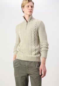 Maglione zippato color crema con design a trecce, tessuto testurizzato, polsini e orlo a costine. Abbinato a pantaloni verde oliva.