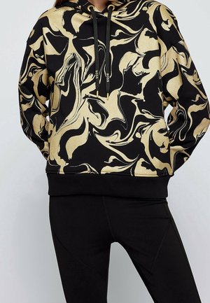 Sort hættetrøje med gyldent abstrakt swirling mønster og sorte snore med gyldne ender, båret med sorte leggings.
