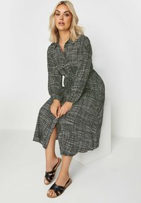 Svart och vit rutigt långärmad klänning med knytmidja, medellång och med sprund på sidan. Bärs med svarta sandaler med korsade remmar på en ljus bakgrund.