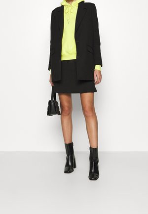 Person, die ein schwarzes Blazer, einen schwarzen Rock, einen neongelben Hoodie, schwarze Absatzstiefeletten trägt und eine kleine schwarze Handtasche hält.