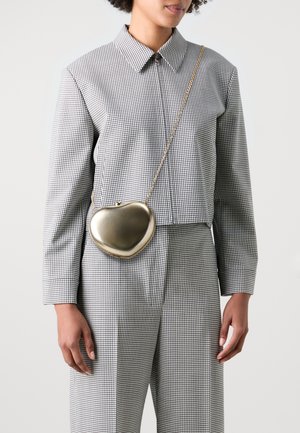 Goldfarbene, herzförmige Handtasche aus Metall mit einer Kettenriemen, getragen über einem schwarz-weiß karierten Hemd und passenden Hosen.