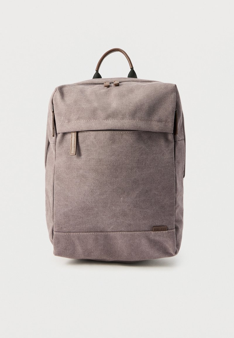 Sac à dos carré en tissu taupe avec une poignée supérieure, une poche avant zippée et un rabat couvrant la fermeture éclair du compartiment principal.