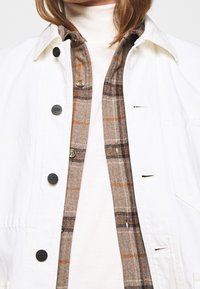 Weiße Denimjacke mit schwarzen Knöpfen, über einem braun-orange karierten Flanellhemd getragen, das über einem cremefarbenen Rollkragenpullover liegt.