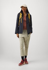 Veste léger navy, pantalon beige avec cordon de serrage, baskets bourgogne et casquette crème ; présente un débardeur gris et des accents de sac �à dos jaunes.