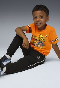 Ein junger Junge sitzt auf dem Boden und trägt ein oranges Puma-T-Shirt mit grafischem Druck, schwarze Puma-Trainingshose und blaue Turnschuhe.