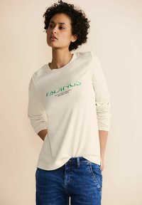 Felpa beige chiaro con maniche lunghe, polsini a coste e scollatura rotonda; presenta un testo verde sul davanti. Abbinata a jeans in denim blu.