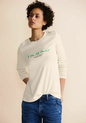 Sweatshirt beige clair à manches longues, avec des poignets côtelés et un col rond ; présente un texte vert sur le devant. Associé à un jean en denim bleu.