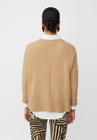 Beige gestrickte Strickjacke mit gerippten Bündchen und Saum, die einen lässigen Schnitt hat. Darunter gemusterte schwarze und beige Hose.