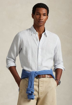 Ριγέ πουκάμισο σε ανοιχτό γαλάζιο και λευκό με κουμπιά, μανίκια γυρισμένα, συνδυασμένο με μπλε πλεκτό πουλόβερ δεμένο στη μέση πάνω από μπεζ παντελόνι.