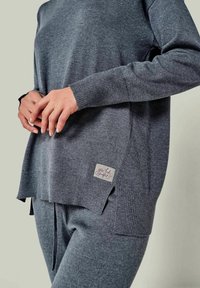 Grauer gestrickter Pullover mit langen Ärmeln und einem seitlichen Schlitz, mit einem kleinen Etikett, auf dem steht "you take perfect." Weiche Textur und lockerer Schnitt.