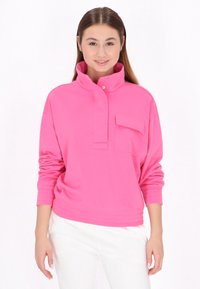 Maglione rosa con colletto alto, chiusura a bottoni a pressione e tasca frontale. Realizzato in un tessuto morbido, presenta polsini e orlo a coste.