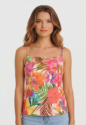 Donna con capelli mossi fino alle spalle, che indossa una canotta senza maniche a fiori colorati e jeans azzurri, in piedi davanti a uno sfondo neutro.