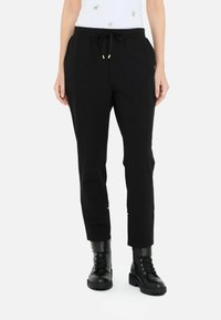 Pantaloni jogger neri con vita regolabile, taglio affusolato e dettagli in tonalità oro. Abbinati a stivali neri con lacci e una maglia chiara.