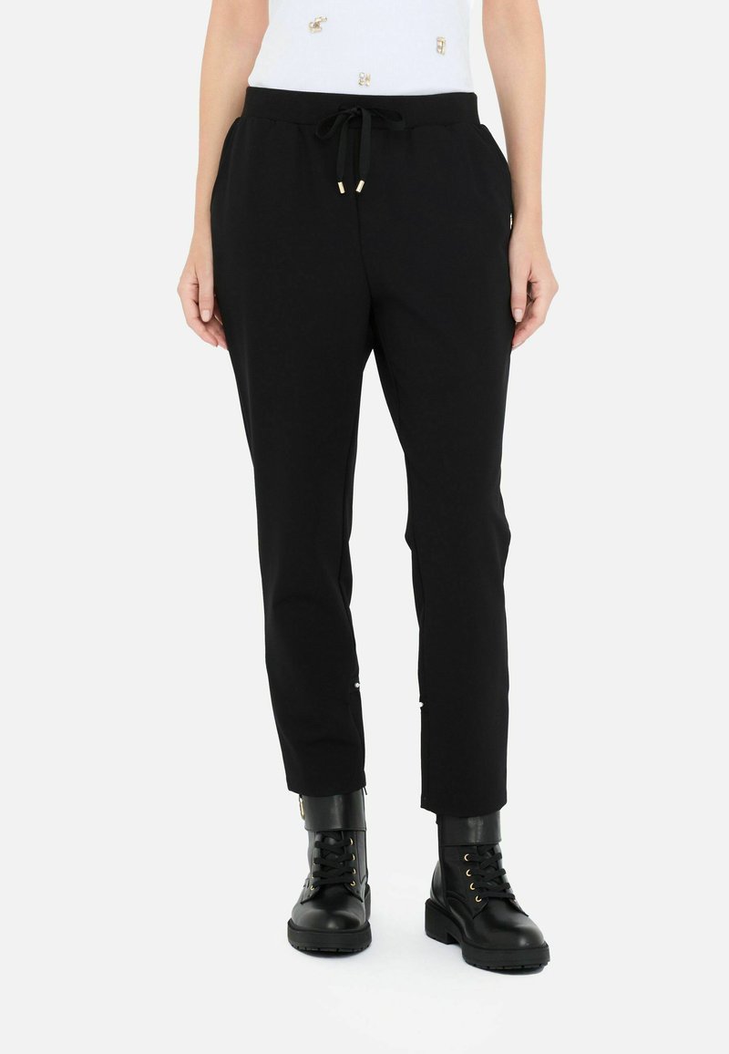 Pantaloni jogger neri con vita regolabile, taglio affusolato e dettagli in tonalità oro. Abbinati a stivali neri con lacci e una maglia chiara.