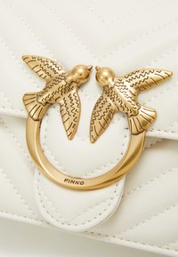 Borsa a tracolla in pelle bianca trapuntata con un fermaglio circolare in tono oro e due decorazioni a forma di uccello. Logo del marchio "PINKO" inciso sul fermaglio.