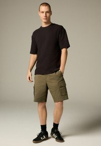 Czarny t-shirt z krótkim rękawem o luźnym kroju, połączony z oliwkowymi szortami cargo z kieszeniami oraz czarnymi trampkami z białymi paskami.