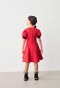 Punainen mekko lyhyillä puffihäivityksillä ja tiukalla bodice-osiolla, jossa on teksturoitu kuvio. Takasaumassa vetoketjukiinnitys. Yhdistetty mustien nilkkasaappaiden kanssa.