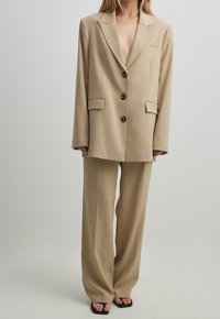 Costume beige sur mesure avec une veste double boutonnage dotée de grands boutons et de deux poches avant, assortie d'un pantalon à jambes larges. Texture douce.