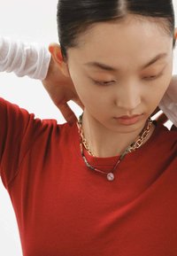 Collares en capas: uno con una cadena dorada, otro con cuentas coloridas texturizadas, que presenta un colgante redondo. Llevado sobre una blusa roja.