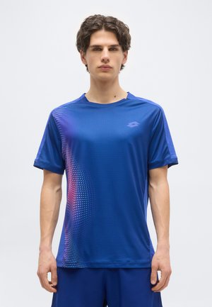 Νεαρός άνδρας που φοράει μπλε αθλητική μπλούζα με ροζ και λευκή διαβαθμισμένη κουκκίδα στην αριστερή πλευρά, στέκεται μπροστά σε ένα απλό ανοιχτό φόντο.