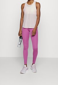 Osoba w różowej koszulce na ramiączkach, magentowych legginsach i jasnoróżowych sneakersach, trzymająca przezroczystą butelkę na wodę z czarną nakrętką i logo.