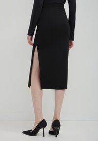 Jupe noire, midi, avec une fente haute sur le côté gauche, en tissu lisse, associée à des escarpins noirs pointus.