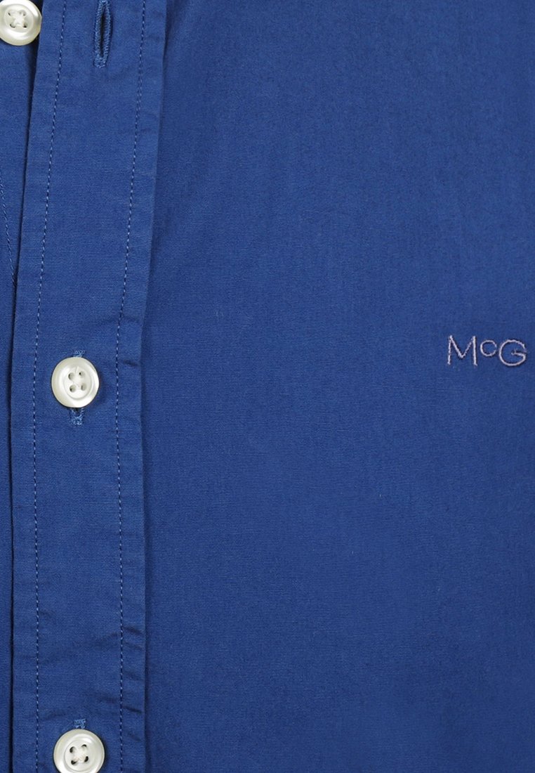 Blaues Hemd mit Knöpfen aus glattem Baumwollstoff. Verfügt über weiße Knöpfe und ein kleines, gesticktes "M&G"-Logo auf der unteren rechten Seite.