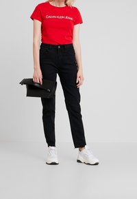Red cotton t-shirt with white logo, black jeans, and white sneakers. Holding a black clutch bag with a flap closure. Simple, casual outfit.