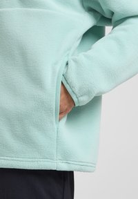 Light blue fleece jacket featuring a side pocket and elastic cuffs, with a soft texture and a rounded hem for comfort.