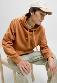 Brun hoodie med snörning och broderad text, i kombination med ljusa denimjeans. Modellen sitter på en trästol mot en enfärgad bakgrund.