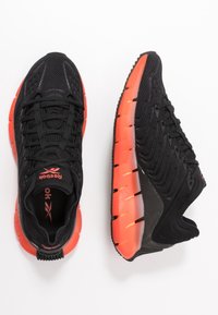 Chaussures de sport noires avec une tige en maille texturée, des accents orange sur la semelle, et un design rembourré pour le confort et le soutien.