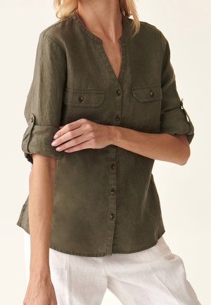 Mujer vistiendo una camisa verde oliva abotonada con las mangas remangadas y pantalones blancos, ajustándose una manga con la mano.