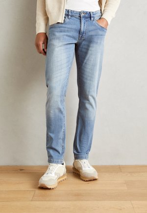 Person trägt hellblaue Slim-Fit-Jeans, beige-weiße Sneaker und eine cremefarbene Strickjacke, steht mit einer Hand in der Tasche auf einem Holzboden.