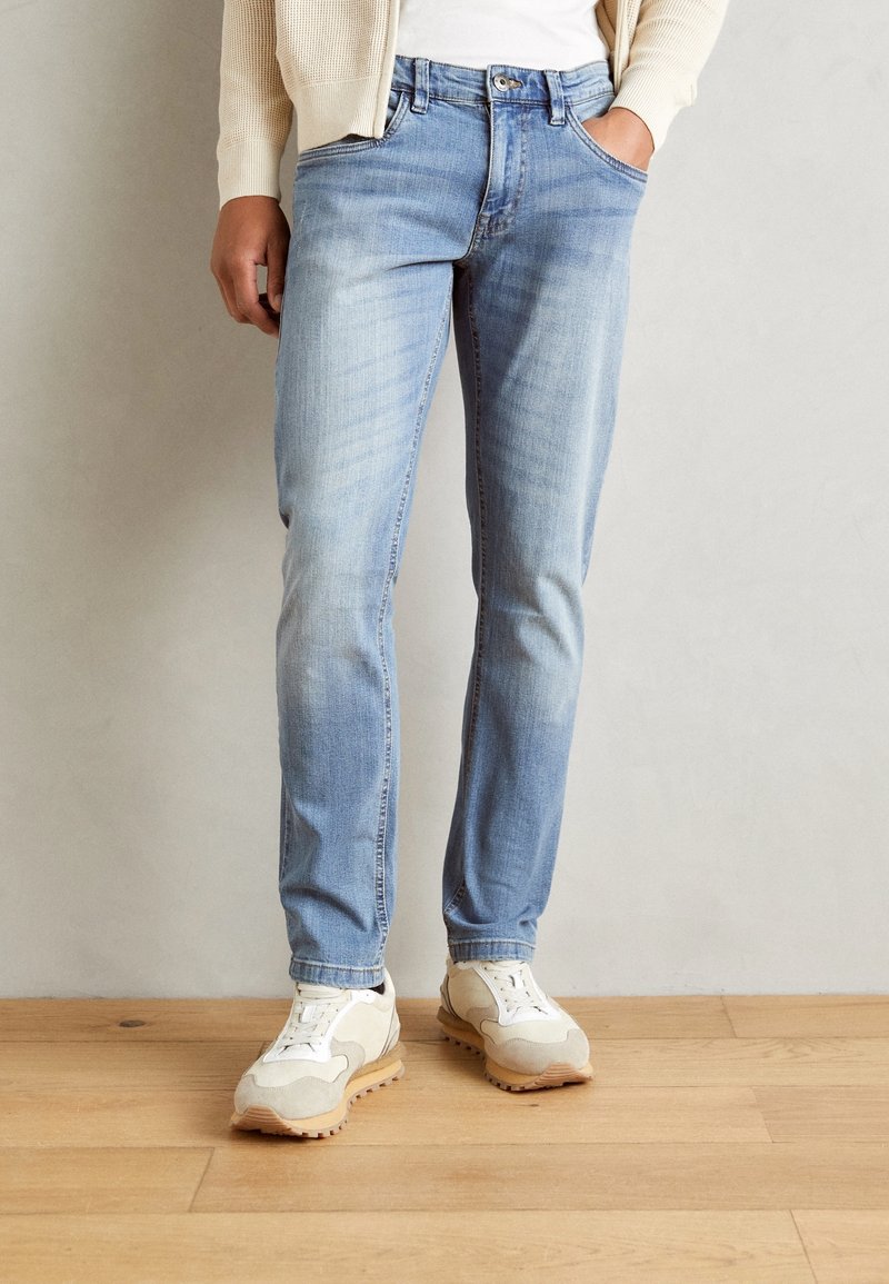 Personne portant un jean slim bleu clair, des baskets beige et blanches, et un cardigan crème, une main dans la poche, debout sur un sol en bois.