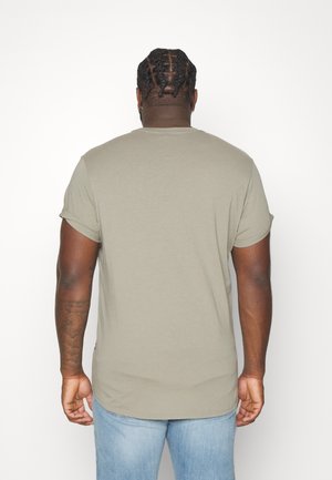 Homme aux cheveux tressés portant une chemise à manches courtes gris clair et un jean bleu, debout de dos sur un fond blanc uni.