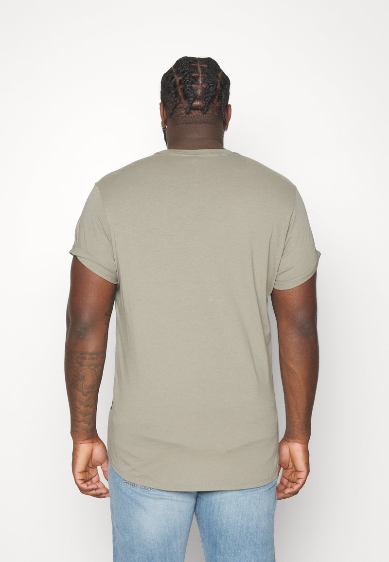 Homme aux cheveux tressés portant une chemise à manches courtes gris clair et un jean bleu, debout de dos sur un fond blanc uni.